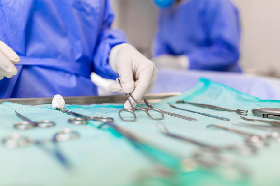 Surgeon in blue surgical gown and gloves selecting an instrument from a sterile tray during C-Section surgery.